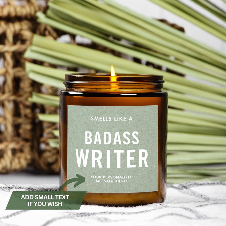May include: A brown glass candle with a green label that reads "Smells like a Badass Writer" and "Your personalized message here!" The candle is lit and the flame is visible.
