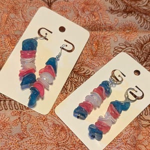 May include: Two pairs of dangle earrings with a colorful design. Each earring features a series of blue, pink, and white beads strung together. The earrings are displayed on white earring cards against a patterned fabric background.