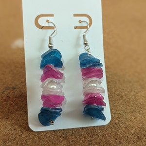 Puede incluir: Un par de pendientes colgantes con un diseño en capas. Los pendientes presentan piezas translúcidas azules, rosas y blancas, intercaladas con una perla. Los pendientes cuelgan de ganchos plateados.