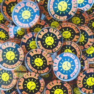 May include: A collection of round buttons with the text "SAD GIRL SUMMER CAMP" in pink and yellow. The buttons feature a yellow sun with a sad face on a blue or black background. The buttons are scattered.