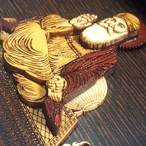 May include: A detailed, carved wooden relief depicting a figure with flowing hair and a beard, in shades of gold, brown, and red. The artwork is mounted on a dark, textured wooden surface, with an ornate border and a small, gold-coloured object.