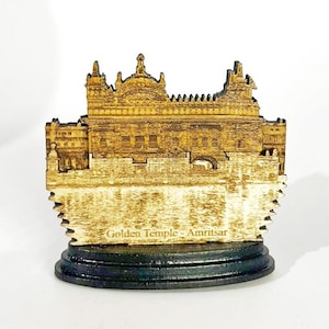 May include: A wooden Golden Temple of Amritsar souvenir. The detailed laser-cut design features the temple's architecture in shades of gold and brown, mounted on a black base. The text "Golden Temple - Amritsar" is visible on the front.