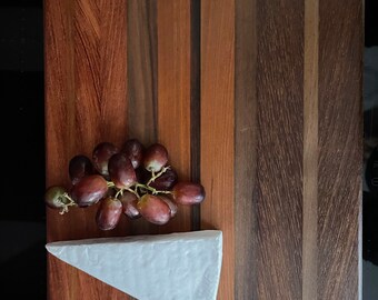 May include: A wooden cutting board with a light brown, dark brown, and reddish brown striped pattern. A wedge of white brie cheese and a bunch of red grapes are on the board.
