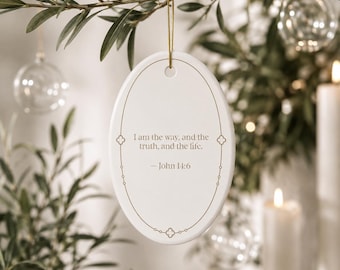 May include: An oval white ceramic ornament featuring a gold-toned border with small clover-like accents. The center displays the text: I am the way, and the truth, and the life. — John 14:6. A thin gold string is attached to the top for hanging.