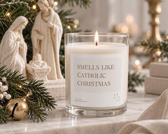 May include: A clear glass candle with a white label featuring the text "SMELLS LIKE CATHOLIC CHRISTMAS" and "11 OZ / 312 G". The candle contains white wax with a single lit wick, positioned in front of a nativity scene figurine.