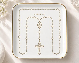 May include: A square white ceramic trinket dish with a gold-colored rim. The center features a gold-colored graphic of a rosary with a crucifix and a central medal. The name AMELIA is printed in gold-colored capital letters above the rosary design.