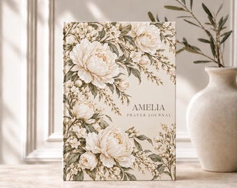 May include: A hardcover prayer journal with a cream-colored cover featuring a floral illustration of white peonies and green leaves. The text AMELIA PRAYER JOURNAL is printed in dark gray lettering on the right side of the front cover.
