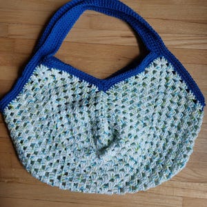 May include: A handmade crochet bag with a blue handle and trim. The body of the bag is white with blue and green flecks. The bag has a rounded bottom and a V-shaped top.