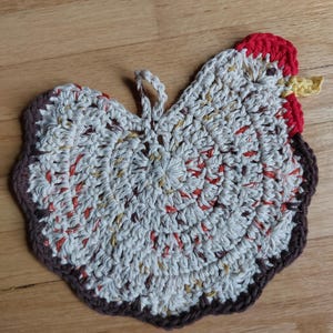 May include: A handmade crochet chicken-shaped hot pad. The pad features a white and speckled body with red, orange, and yellow accents. The edges are brown, and the comb is red. A loop is attached for hanging. Kitchen decor.