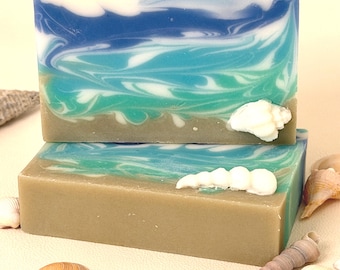 Puede incluir: Dos barras rectangulares de jabón artesanal con un diseño de playa. El jabón presenta capas de azul, turquesa y blanco, que se asemejan a las olas del océano, sobre una base beige. Conchas marinas dispersas alrededor de las barras de jabón.