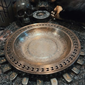 May include: A silver-toned, ornate plate with a decorative rim, surrounded by small, rectangular stones with gold-colored symbols. The plate sits on a dark, speckled surface, with skulls and other gothic decor in the background.