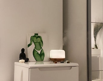 May include: White chest of drawers with three drawers, a white diffuser with a wooden base, a small black statue, and a green abstract painting of a person's torso.
