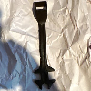以下が含まれることがあります： 黒い鋳鉄製の工具。長く細いハンドル、上部の長方形の開口部、そして下部には特定の機械部品を掴んだり回転させたりするための複数の突起を持つ特殊なヘッドが備わっています。