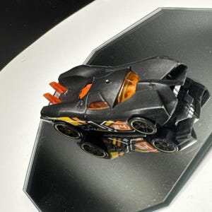 May include: A black toy race car featuring orange windows, orange rear fins, and yellow and orange side decals with the number 24 printed on the side. The vehicle has black wheels with yellow rims and a low-profile, aerodynamic body design.