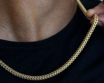 May include: A gold chain necklace with a curb link style. The chain is worn by a person and is visible against a black shirt.