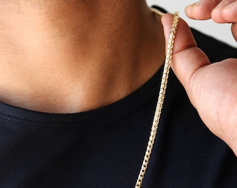 May include: A gold chain necklace with a classic curb link design. The chain is worn by a person with a black shirt.