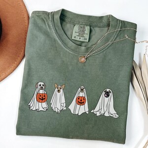 Puede incluir: Camiseta verde oliva Comfort Colors con un gr&aacute;fico de cuatro perros disfrazados de fantasmas, cada uno sosteniendo una calabaza naranja. Tambi&eacute;n se ven un collar dorado y un sombrero marr&oacute;n. La camiseta es talla peque&ntilde;a.