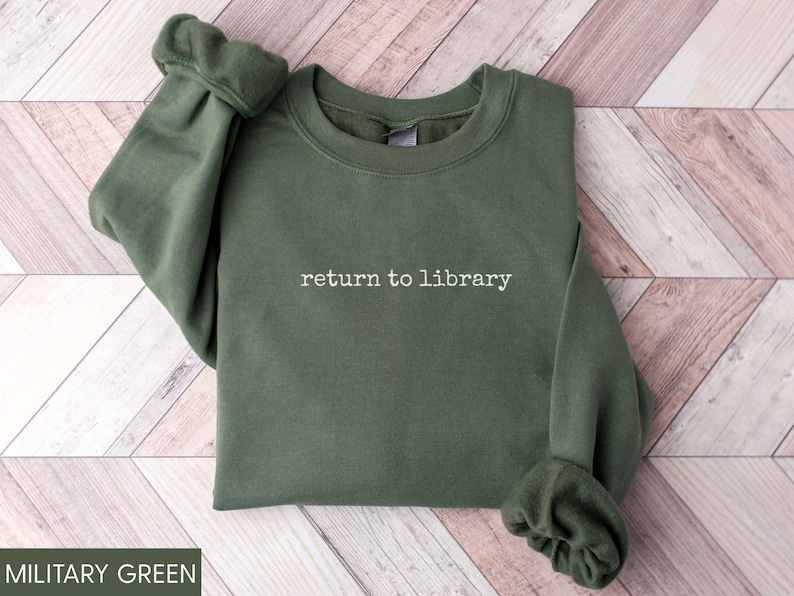 May include: Military green sweatshirt with the phrase "return to library" in white script. The long-sleeved top is made of a soft material and is folded neatly. The sleeves are cuffed at the wrists. The sweatshirt is displayed on a light brown and white wooden surface.