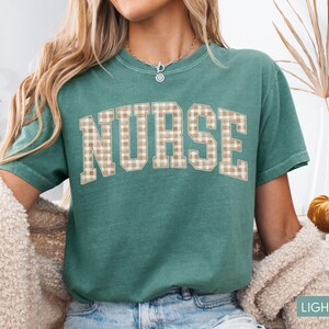 May include: A light green t-shirt with the word "NURSE" in large, tan plaid letters. The shirt has short sleeves and a crew neck. The text "LIGHT GREEN" is at the bottom right corner of the image.