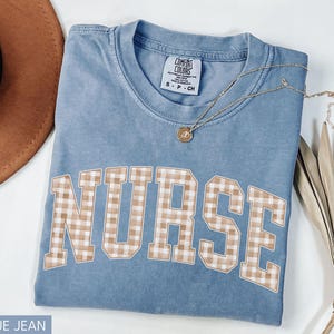 May include: Blue jean colored t-shirt with the word "NURSE" in a tan and white gingham pattern. A gold necklace with a pendant is on the shirt. The shirt is folded on a white surface.
