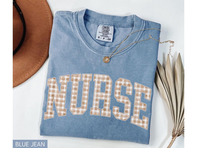 May include: Blue jean colored t-shirt with the word "NURSE" in a tan and white gingham pattern. A gold necklace with a pendant is on the shirt. The shirt is folded on a white surface.