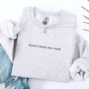 May include: A light gray sweatshirt with the text "that's what she read" in black. A silver necklace with a small pendant rests on the sweatshirt. The sweatshirt is paired with blue jeans.