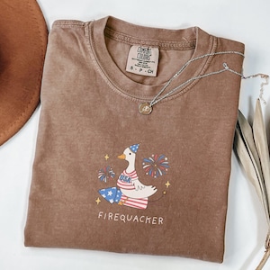 Op de afbeelding: Een bruin T-shirt met een cartoon-gans die een feestmuts draagt en vuurwerk vasthoudt. De tekst "Firequacker" is onder de gans gedrukt. Het shirt heeft een verweerde look en is gemaakt door Comfort Colors.