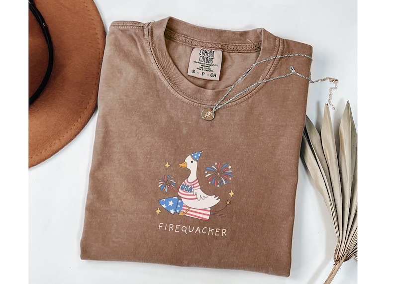 Op de afbeelding: Een bruin T-shirt met een cartoon-gans die een feestmuts draagt en vuurwerk vasthoudt. De tekst "Firequacker" is onder de gans gedrukt. Het shirt heeft een verweerde look en is gemaakt door Comfort Colors.