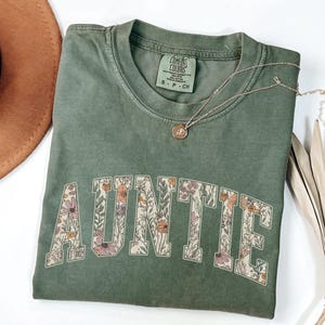 May include: A green t-shirt with the word "AUNTIE" printed in white letters outlined with a floral pattern. The shirt is made of a soft, comfortable material and has a relaxed fit.