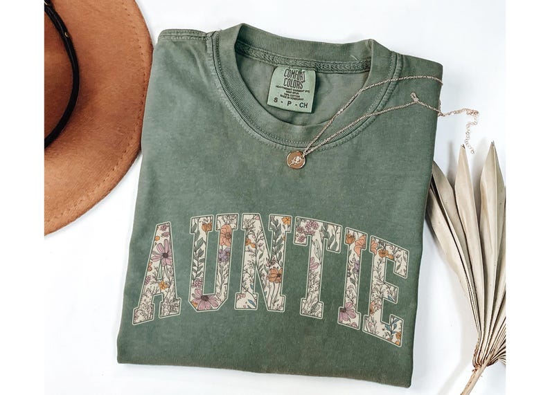 May include: A green t-shirt with the word "AUNTIE" printed in white letters outlined with a floral pattern. The shirt is made of a soft, comfortable material and has a relaxed fit.