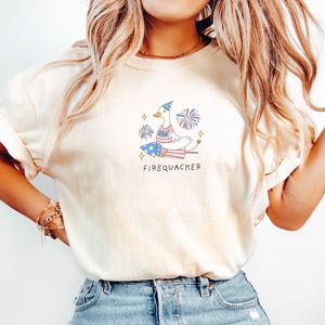 Op de afbeelding: Een wit T-shirt met een afbeelding van een cartoon eend die een rood, wit en blauw hoedje draagt en op een vuurwerk zit. De tekst "Firequacker" staat onder de afbeelding.