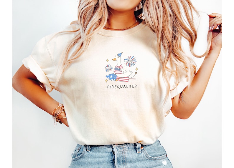 Op de afbeelding: Een wit T-shirt met een afbeelding van een cartoon eend die een rood, wit en blauw hoedje draagt en op een vuurwerk zit. De tekst "Firequacker" staat onder de afbeelding.