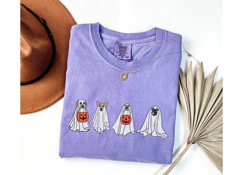 Puede incluir: Una camiseta lila Comfort Colors con cuatro perros disfrazados de fantasmas, cada uno con una calabaza. Un collar dorado est&aacute; sobre la camiseta. Un sombrero marr&oacute;n y una hoja de palma seca est&aacute;n en el fondo.
