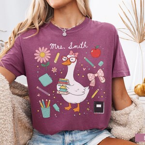 以下が含まれることがあります： ベリー色のTシャツ。メガネをかけ、本を抱えたガチョウが学校をテーマにしたイラストに囲まれた、気まぐれなグラフィックが特徴です。「Mrs. Smith」の文字がガチョウの上にあります。