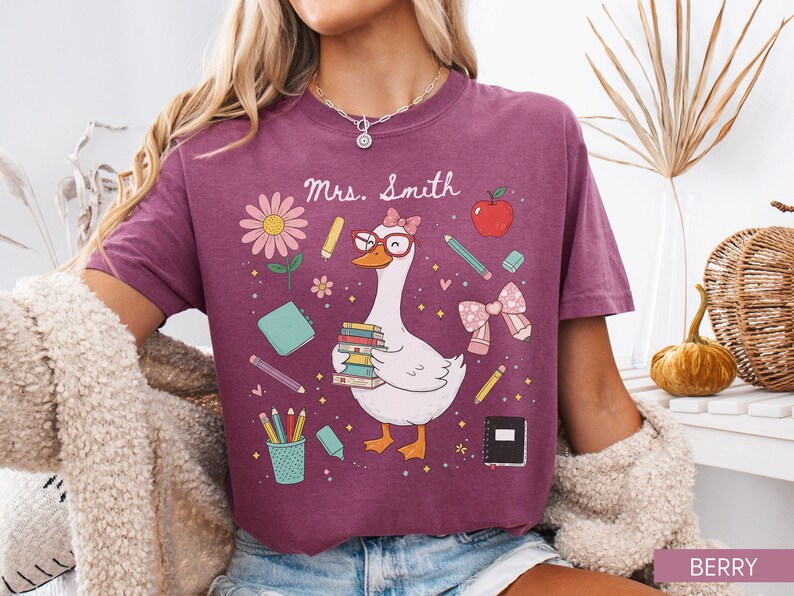 以下が含まれることがあります： ベリー色のTシャツ。メガネをかけ、本を抱えたガチョウが学校をテーマにしたイラストに囲まれた、気まぐれなグラフィックが特徴です。「Mrs. Smith」の文字がガチョウの上にあります。