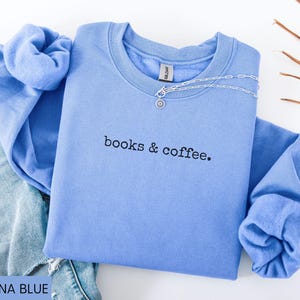 May include: A Carolina blue sweatshirt with the text "books & coffee" in black. A silver necklace with a pendant is draped across the neckline. The sweatshirt is folded on top of a pair of blue jeans.