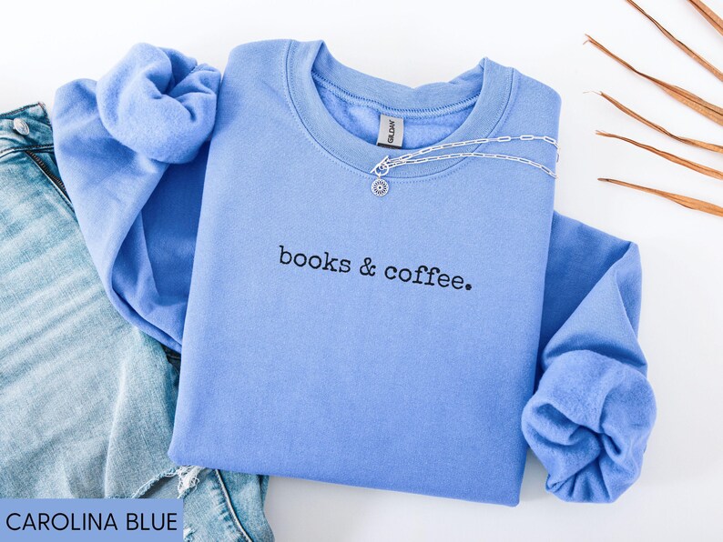 May include: A Carolina blue sweatshirt with the text "books & coffee" in black. A silver necklace with a pendant is draped across the neckline. The sweatshirt is folded on top of a pair of blue jeans.