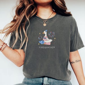Op de afbeelding: Een donkergrijs T-shirt met een cartoonillustratie van een eend die een feestmuts draagt en vuurwerk vasthoudt. De tekst "FIREQUACKER" is onder de eend gedrukt.