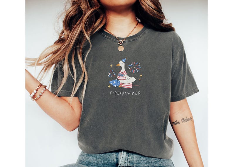 Op de afbeelding: Een donkergrijs T-shirt met een cartoonillustratie van een eend die een feestmuts draagt en vuurwerk vasthoudt. De tekst "FIREQUACKER" is onder de eend gedrukt.