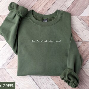 May include: Military green sweatshirt with the phrase "that's what she read" embroidered in white. The long-sleeved top is made of a soft material and is folded neatly. The sleeves have a gathered cuff detail.