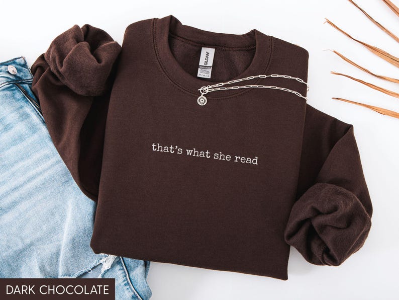 May include: A dark chocolate-colored sweatshirt with the embroidered text "that's what she read." A silver necklace with a pendant is draped across the front. The sweatshirt is paired with blue jeans.