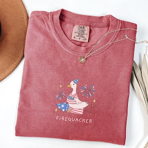 Op de afbeelding: Een rood T-shirt met een cartoonillustratie van een gans die een feestmuts draagt en een vuurwerk vasthoudt. De gans draagt een blauw-wit gestreept shirt met sterren en de tekst "USA". De tekst "Firequacker" staat onder de gans. Het T-shirt heeft een verweerde look en is gemaakt door "Comfy Colors".