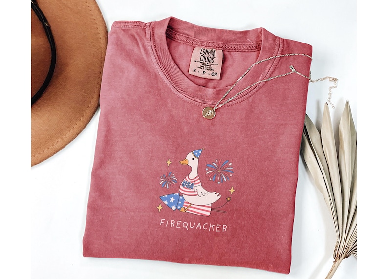 Op de afbeelding: Een rood T-shirt met een cartoonillustratie van een gans die een feestmuts draagt en een vuurwerk vasthoudt. De gans draagt een blauw-wit gestreept shirt met sterren en de tekst "USA". De tekst "Firequacker" staat onder de gans. Het T-shirt heeft een verweerde look en is gemaakt door "Comfy Colors".