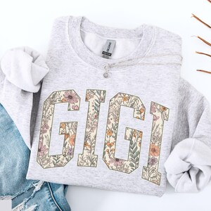 May include: Pale grey sweatshirt featuring the name "GIGI" in large, floral-patterned letters. The letters are filled with a design of flowers and leaves in shades of pink, orange, and green. The sweatshirt is paired with blue jeans.