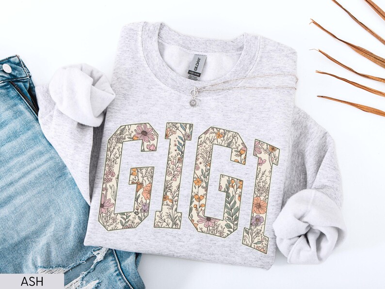 May include: Pale grey sweatshirt featuring the name "GIGI" in large, floral-patterned letters. The letters are filled with a design of flowers and leaves in shades of pink, orange, and green. The sweatshirt is paired with blue jeans.