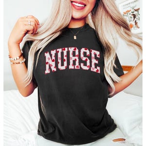Pode incluir: Uma camiseta preta com um contorno branco da palavra "NURSE" em cora&ccedil;&otilde;es vermelhos e rosa.