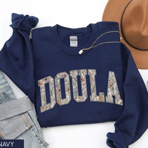 May include: Navy blue sweatshirt with the word "DOULA" in large, floral-filled letters. The sweatshirt is folded, with a pair of jeans, a brown hat, and a gold necklace visible. A small plant is also in the image.