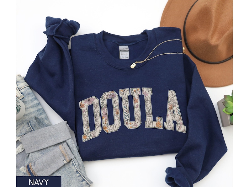 May include: Navy blue sweatshirt with the word "DOULA" in large, floral-filled letters. The sweatshirt is folded, with a pair of jeans, a brown hat, and a gold necklace visible. A small plant is also in the image.