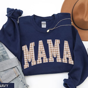 May include: Navy blue sweatshirt with the word "MAMA" in a tan and white gingham pattern. A gold necklace and a brown hat are also visible. The sweatshirt is folded on top of a pair of blue jeans.