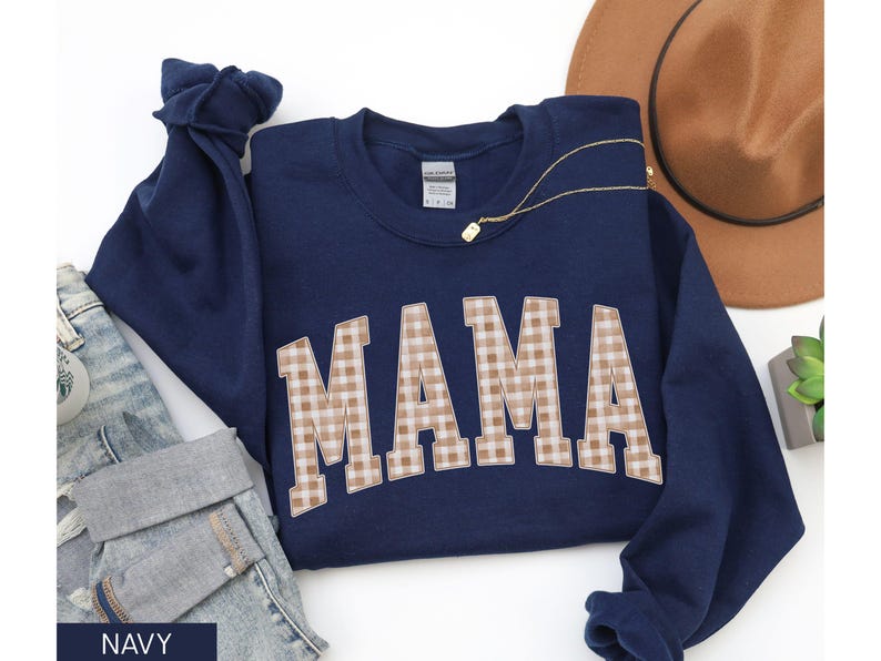 May include: Navy blue sweatshirt with the word "MAMA" in a tan and white gingham pattern. A gold necklace and a brown hat are also visible. The sweatshirt is folded on top of a pair of blue jeans.